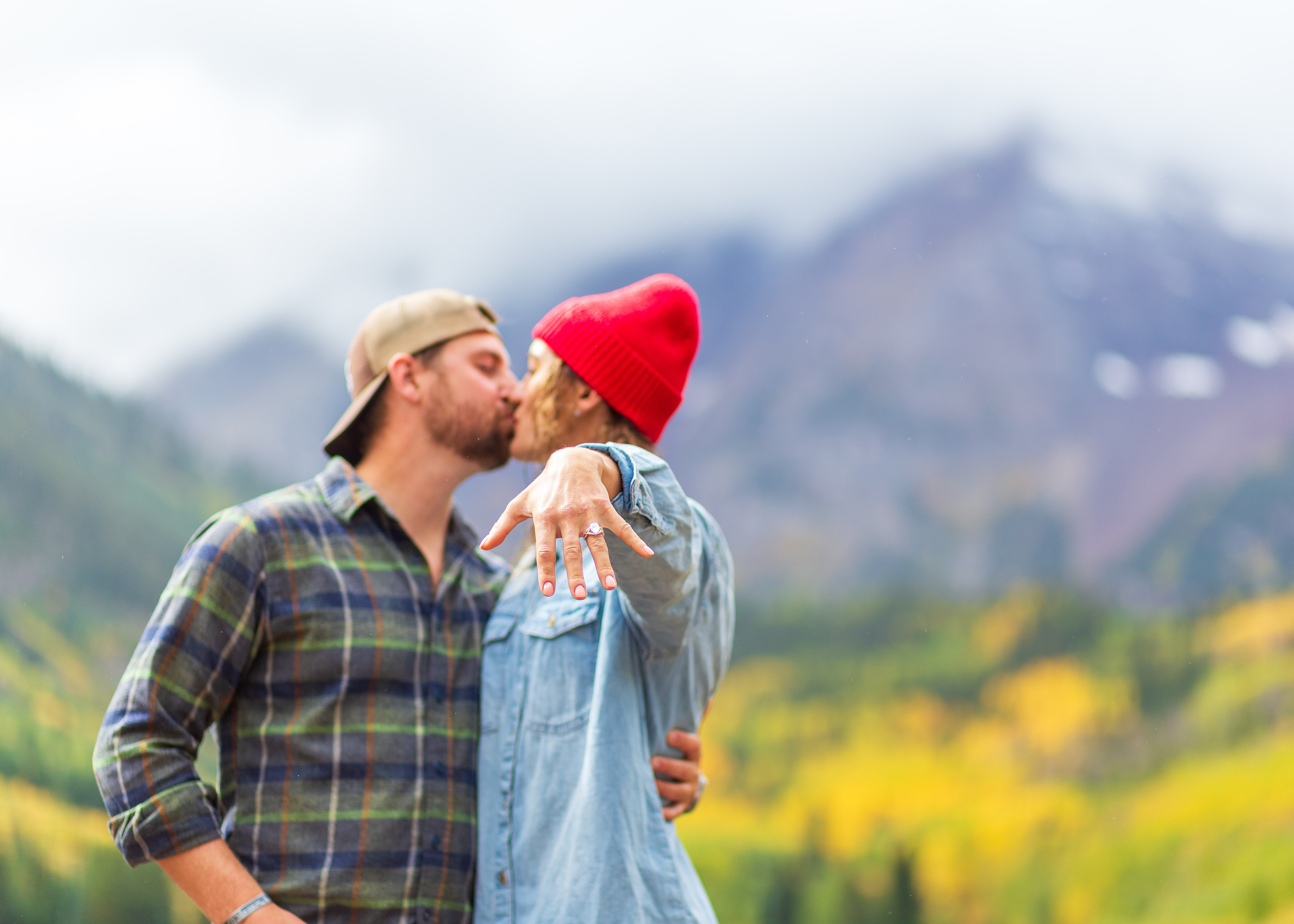 Ren\u00e9e and Alexander kissing in front of mountains with autumn foliage, showing engagement ring